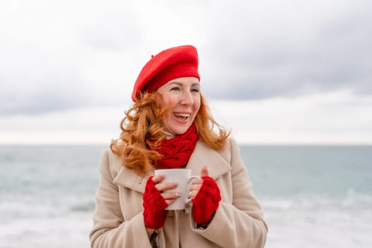 Woman beach in red beret scarf and mitts holding a cup of tea in his hands. Depicting beach relaxation and cozy attire. Walks by the sea.