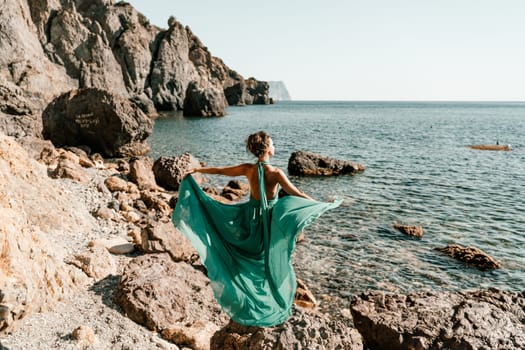 Woman green dress sea. Woman in a long mint dress posing on a beach with rocks on sunny day. Girl on the nature on blue sky background