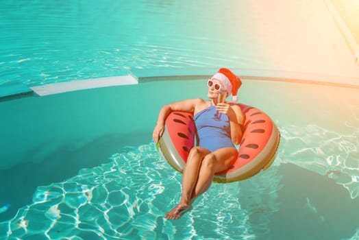 A happy woman in a blue bikini, a red and white Santa hat and sunglasses poses in the pool in an inflatable circle with a watermelon pattern, holding a glass of champagne in her hands. Christmas holidays concept