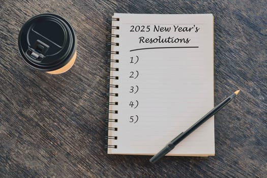 2025 New Year's Resolutions text on notepad with pen and disposable coffee cup on wood desk.