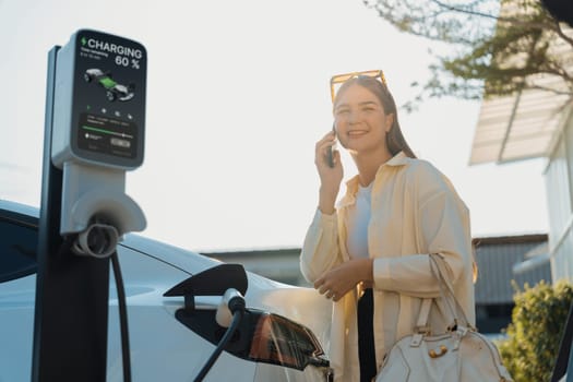 Young woman holding shopping bag talking on the phone while recharging EV car battery from charging station at city mall parking lot. Modern woman go shopping by eco car. Expedient