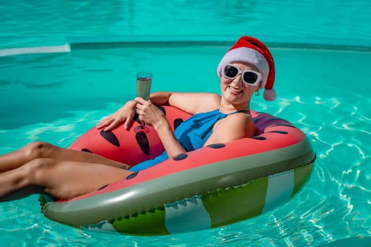 A happy woman in a blue bikini, a red and white Santa hat and sunglasses poses in the pool in an inflatable circle with a watermelon pattern, holding a glass of champagne in her hands. Christmas holidays concept