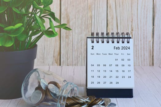 February 2024 desk calendar with stack of coins and potted plant on wooden desk. Business and finance deadline concept.
