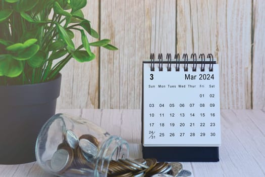 March 2024 desk calendar with stack of coins and potted plant on wooden desk. Business and finance deadline concept.