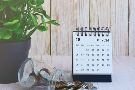 October 2024 desk calendar with stack of coins and potted plant on wooden desk. Business and finance deadline concept.