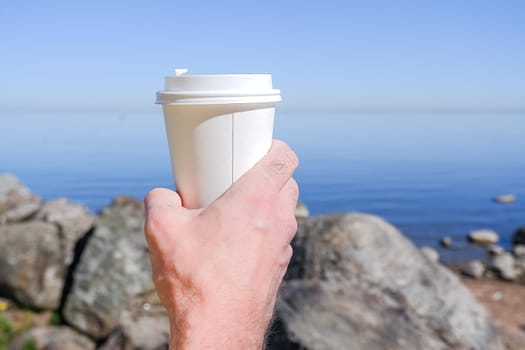 Morning paper coffee cup with tasty hot coffee in men hand outdoors on sea background.