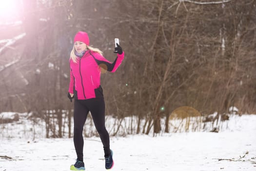 Athletic woman taking selfie with her smartphone and wearing headphones while jogging on a bridge during winter snowy day. Healthy lifestyle, winter fitness, cold weather. High quality photo