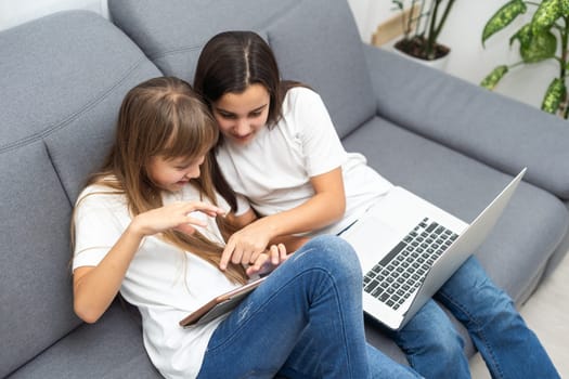 Young twins using a laptop and a tablet sitting on a couch in the living room. High quality photo