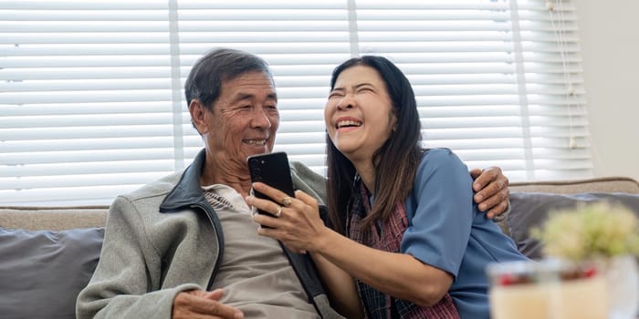 Happy elderly asian couple using smartphone sit on sofa doing ecommerce shopping online on website at home.