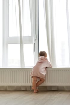 Little child girl standing at window and looking outside. Back view.