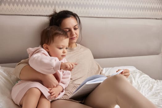 beautiful mother reading book to her cute little baby daughter on bed. family spending time together