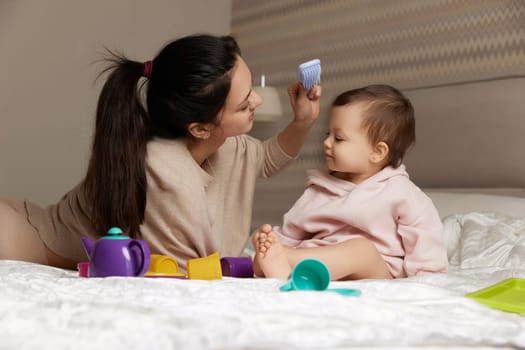 attractive mother is combing her cute little daughter hair on bed