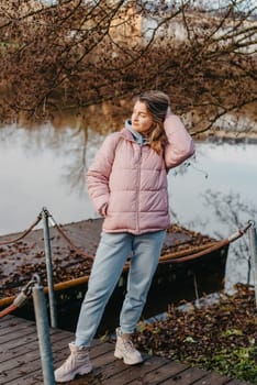 A young woman standing at the shore looking at the river in autumn sunny day. Street view, copy space for text, travel photo. Happy tourist woman on the bank of the river in autumn in warm clothes. Tourists enjoy their vacation, winter season. Romantic look and travel concept. A joyful mood in a Caucasian girl. Winter Wonderland: Enchanting Girl by the Riverside in Autumn.