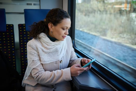 Attractive young adult woman using smart phone while enjoying her business travel by train, sitting on a seat by window overlooking downtown. People. Lifestyle. Public railroad transportation concept