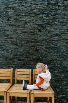 Little girl sits on a row of wooden chairs against the wall and looks at her shoes. Side view. High quality photo