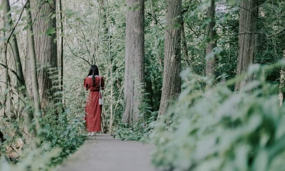 Portrait of one beautiful young Caucasian brunette girl from the back in a long red dress walking along a wooden path in a green forest on a spring day, close-up view from below.