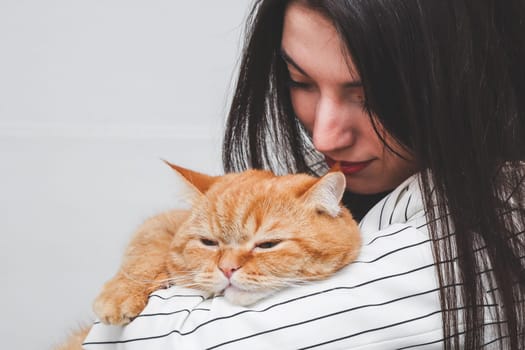 Portrait of one beautiful young Caucasian brunette with long and straight hair in a classic striped black and white jacket holding in her arms a red purebred cat with a satisfied emotion lying on her arm and hanging her paw against the background of a white wall in the room, close-up side view.