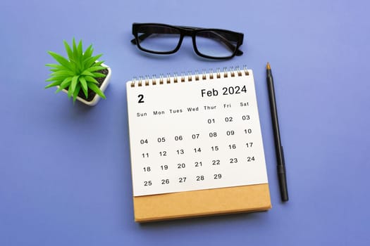 February 2024 desk calendar with potted plant, pen and reading glasses on blue background.