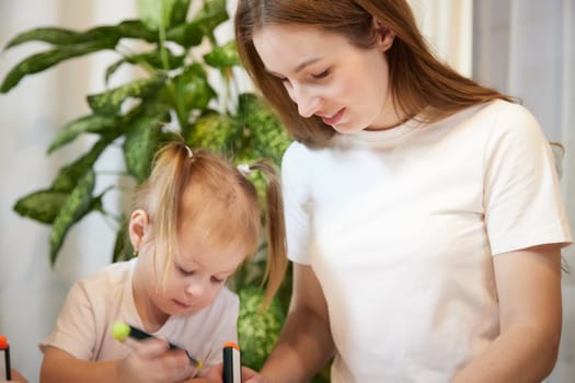 Young mother or babysitter, little daughter drawing at table in room. Painting, doing homework. Family enjoying leisure at home