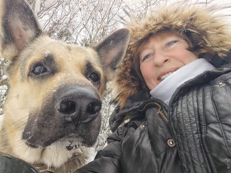 Adult girl with shepherd dog taking selfies in winter forest. Middle aged woman and big shepherd dog on nature in cold day. Friendship, love, communication, fun, hugs