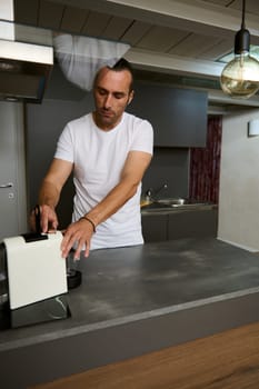 Hispanic attractive young man inserting a capsule with grinded coffee inside a coffee machine for home use, preparing freshly brewed espresso for breakfast in the morning, standing at kitchen counter