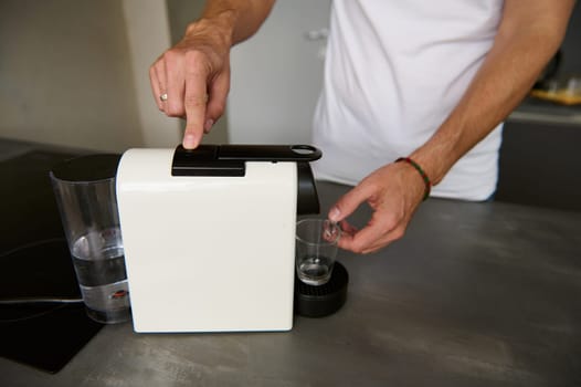 Selective focus on a capsule coffee machine for home use and man preparing fresh morning coffee for breakfast, standing by kitchen counter. Italian lifestyle and culture.