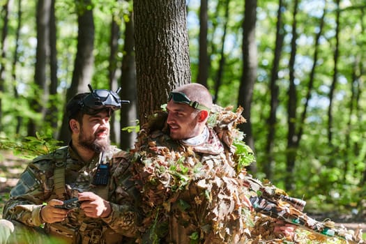 A skilled sniper and a soldier operating a drone with VR goggles strategize and observe the military action while concealed in the forest.