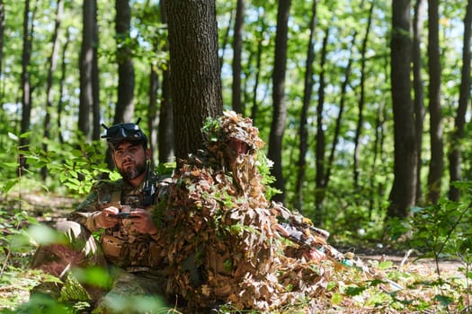 A skilled sniper and a soldier operating a drone with VR goggles strategize and observe the military action while concealed in the forest.