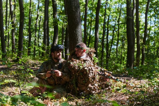 A skilled sniper and a soldier operating a drone with VR goggles strategize and observe the military action while concealed in the forest.