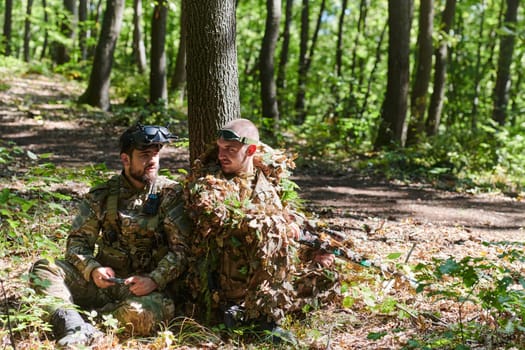 A skilled sniper and a soldier operating a drone with VR goggles strategize and observe the military action while concealed in the forest.