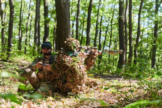 A skilled sniper and a soldier operating a drone with VR goggles strategize and observe the military action while concealed in the forest.