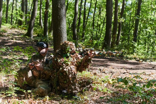 A skilled sniper and a soldier operating a drone with VR goggles strategize and observe the military action while concealed in the forest.