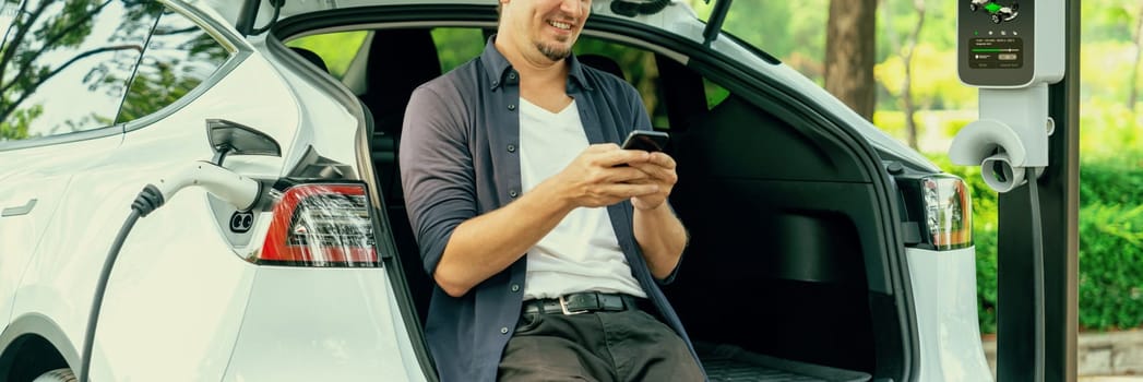 Man using smartphone online banking application to pay for electric car battery charging from EV charging station during vacation road trip at national park or summer forest. Panorama Exalt