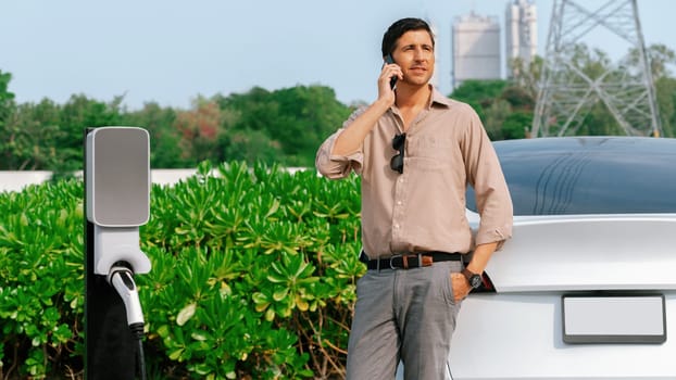 Man talking on the phone while recharge EV car battery at charging station connected to power grid tower electrical as electrical industry for eco friendly car utilization. Panorama Expedient