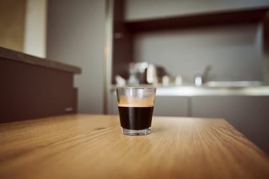 Selective focus on a glass cup of freshly brewed espresso coffee standing on a wooden kitchen table. Still life. Copy advertising space. The concept of caffeinated drink