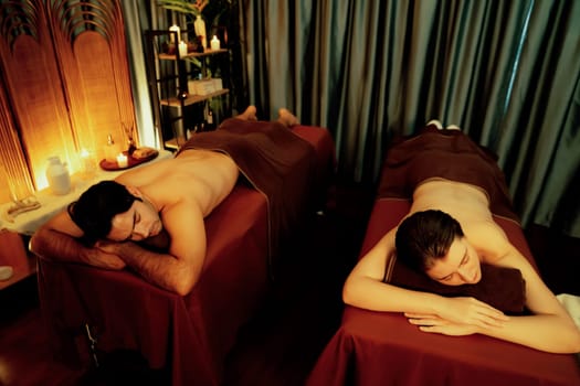 Caucasian couple customer enjoying relaxing anti-stress spa massage and pampering with beauty skin recreation leisure in warm candle lighting ambient salon spa at luxury resort or hotel. Quiescent