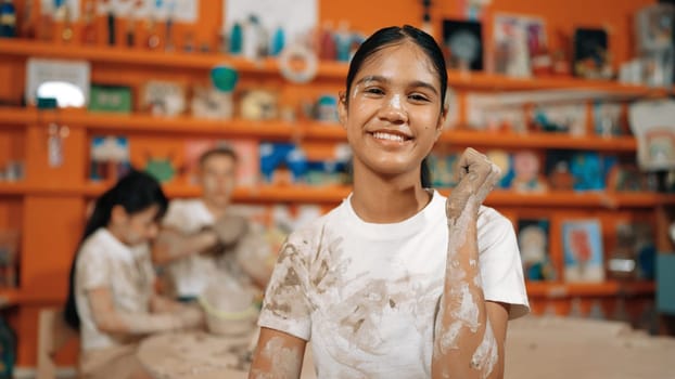 Happy caucasian girl pose at camera while diverse children modeling clay behind. Cute student wearing dirty shirt while looking at camera at workshop in art lesson. Blurring background. Edification.