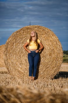 A young beautiful girl in a field stands near a sheaf. Vacation in the village. Young woman in yellow top and jeans