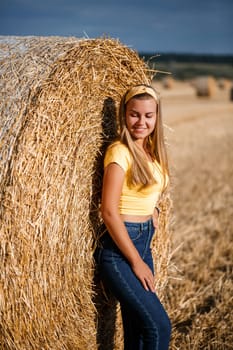 A young beautiful girl in a field stands near a sheaf. Vacation in the village. Young woman in yellow top and jeans