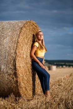 A young beautiful blonde stands on a mown wheat field near a huge sheaf of hay, enjoying nature. Nature in the village