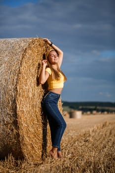 A young beautiful blonde stands on a mown wheat field near a huge sheaf of hay, enjoying nature. Nature in the village