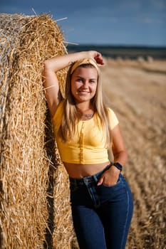 A young beautiful blonde stands on a mown wheat field near a huge sheaf of hay, enjoying nature. Nature in the village