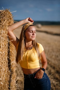 A young beautiful girl in a field stands near a sheaf. Vacation in the village. Young woman in yellow top and jeans