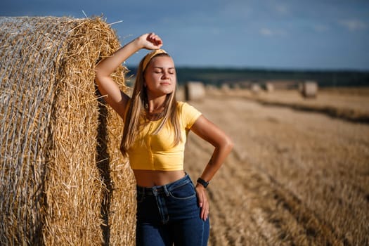 A young beautiful blonde stands on a mown wheat field near a huge sheaf of hay, enjoying nature. Nature in the village