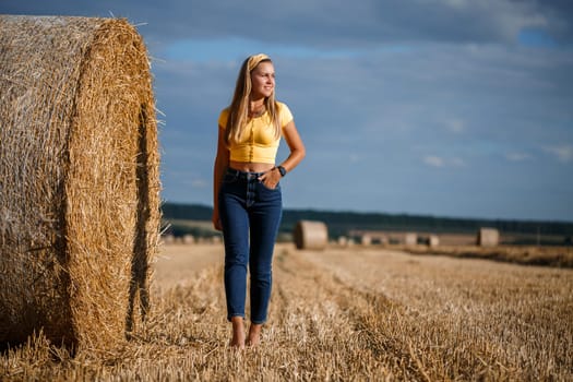 A young beautiful blonde stands on a mown wheat field near a huge sheaf of hay, enjoying nature. Nature in the village