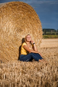 Beautiful young woman near a sheaf of hay in a field. Holidays in the village, a girl enjoying nature in a mown field on a sunny day