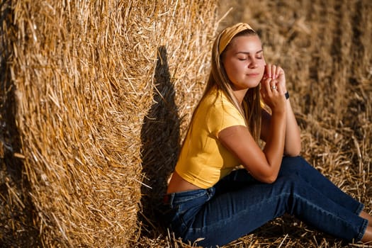 Beautiful young woman near a sheaf of hay in a field. Holidays in the village, a girl enjoying nature in a mown field on a sunny day