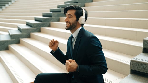 Project manager listening and enjoy music from headphone while sitting at stairs. Professional businessman wearing suit outfit while dancing and moving to music. Happy man listen funny song. Exultant.