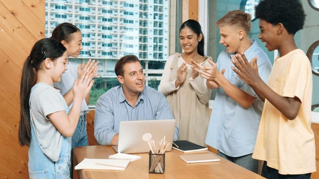 Caucasian teacher and multicultural students clapping hand or putting the hand in the air together to celebrate for successful project with laptop and equipment placed on table in class. Edification.