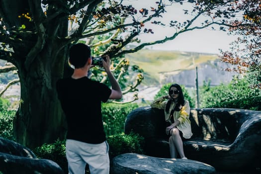 A young man standing from the back takes a photograph of a sitting girl on a wooden bench against the backdrop of rocks and beautiful nature on a sunny summer day in the botanical garden of wonders in France, close-up side view with depth of field.